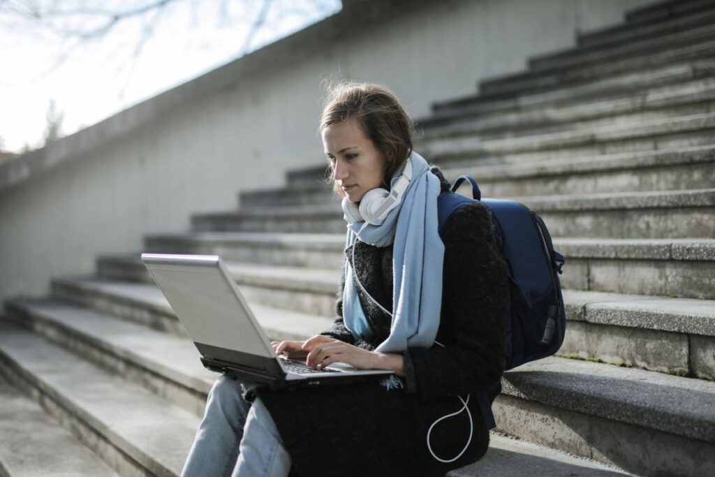 Student sitting on outdoor steps with a backpack and headphones, working on a laptop between classes