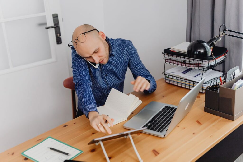 Person at a home desk juggling a phone call, a laptop and a paper notebook while managing admin tasks for clients